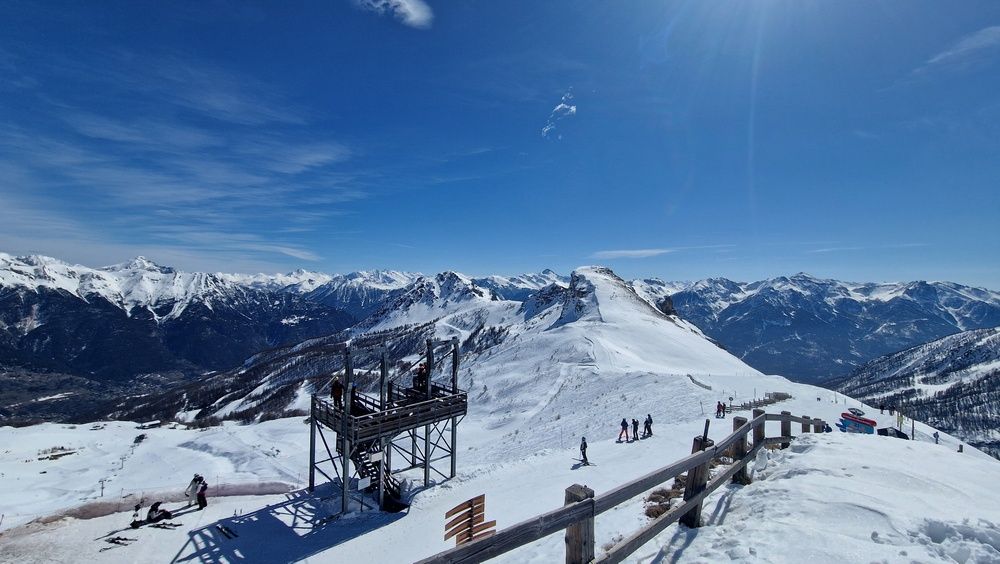 Skiers at a mountain-top viewing platform with a panorama of snow-capped Alpine peaks