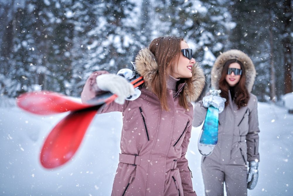 Two skiers in stylish ski suits and sunglasses carrying skis through falling snow